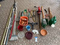 Top-down view of the hedge trimmer, fishing rods, pruning shears, flower pots, watering cans, broom, ropes, and other small gardening tools arranged on carpet.