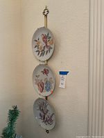 Full view of the three hand-painted Nora Fenton plates mounted vertically on the brass wall hanger against a beige wall.