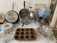 Full assortment of non-matching cookware and gadgets including frying pans, bakeware, colander, salad spinner and more shown on table.