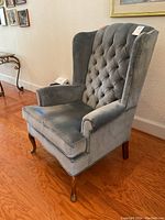 Side angled view of the blue tufted Queen Anne wingback chair showing wooden cabriole leg and velvet upholstery.