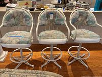 Front view of three matching white metal swivel barstools with pastel abstract upholstery on cushions, placed side by side.