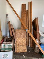 Photo showing two wide 2x12 boards and multiple 2x4 planks leaning vertically against a wall, surrounded by some boxes and materials.
