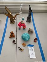 Top-down view of all 10 whimsical figurines on a white table including the brass duck, glass bird, clown doll, Statue of Liberty, and various small figures with a blue round container.