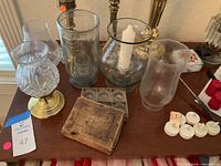 Photo of assorted vintage candle holders made of brass and glass, glass hurricane candle covers, small box of tealight candles, and some pillar candles assorted on wooden table surface