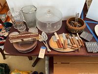 Wide view of all vintage serving items arranged on table including glass pitcher, cake plate with etched dome, cutting board with knives and utensils, napkin rings, nut set, glasses, plates, and coffee stirrers.