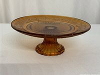 Side angle photo showing the amber glass cake stand with intricate pattern around the edges and fluted pedestal base