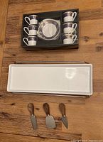 Set of 6 espresso cups and saucers in box, white with blue trim; one cup has broken handle. Below, long white rectangular ceramic dish with metal holder and three wooden handle cheese knives.