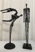Front view of two twisted metal wire sculptures, one female dancing figure and one tall male figure.