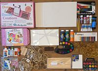 Overview of art supplies including watercolor paint set, various acrylic paint bottles, paper craft kits, natural wood chip decor and gold metal wire decor on table.