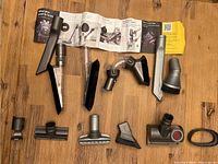 Dyson V11 vacuum cleaner attachments laid out on wooden floor including various crevice tools, brushes, adapters, and instruction manual.