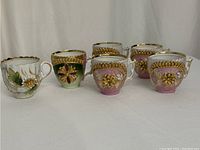 Photo showing six ornately decorated porcelain cups with gold trim and floral detailing in pink, green, and white colors.