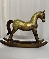 Side view of decorative rocking horse showing detailed gold-tone embossing with floral patterns and dark wood base and tail.