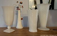 Four white milk glass vases arranged side by side showing different shapes and textured designs; one vase holds a wire floral bouquet made with antique buttons.