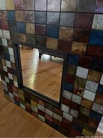 Front and angled view showing the mirror and colorful mosaic tile frame with varied earthy and metallic tones, positioned on a wooden floor.