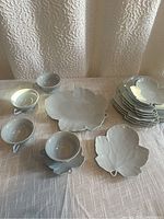 Overall view of 4 large leaf-shaped plates, 4 smaller salad plates stacked, 4 cups, and 4 leaf-shaped saucers arranged on tablecloth.