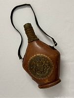 Front angle view of vintage leather-wrapped flask with strap and embossed emblem