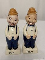 Front view of both vintage ceramic salt and pepper shakers named Pop and Ray, showing detailed facial expressions, attire, and labeling on base.