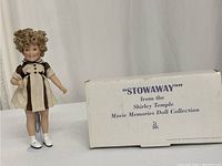 Shirley Temple 8 inch doll standing beside the original white box with the label 'Stowaway' from the Movie Memories Doll Collection.