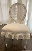 View of front of white wood chair with oval rattan back and cream removable seat cover with ruffles.