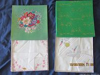 Two green decorative boxes each containing folded white embroidered handkerchiefs seen from above. One box lid has a colorful floral design, the other has gold text patterning. Handkerchiefs are cotton with embroidery details visible.