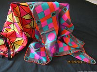 Two silk scarves draped on a black background showing vibrant geometric patterns and bright colors of pink, orange, yellow, purple, turquoise, and brown.