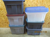 Eight stacked large plastic storage containers with lids in various colors against a wooden background. Containers are gray and white; lids are blue, red, and green.
