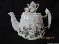 Front view of large white ceramic teapot with floral and insect decoration and mini tea set on lid