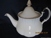 Side view of the white porcelain Royal Albert teapot showing the silver trim, curved handle with silver accent, and the repaired pointed finial on the lid.