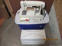 Vintage Singer sewing machine on a blue plastic tub with foot pedal and plastic storage case for accessories, plus a hard white carrying case placed in front.