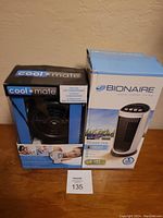 Two boxed tabletop fans on wooden surface against beige wall: Cool Mate personal fan in black, Bionaire tower fan in white/black.