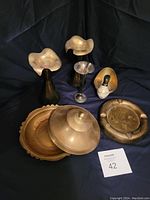 Three brass vases, covered dish with lid, duck candy dish, and brass ashtray set against a black background.