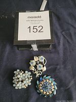 Top-down photo of three brooches on dark fabric with auction lot number card visible