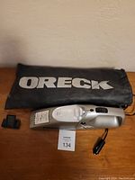 ORECK handheld vacuum, attachment nozzle, and storage bag displayed on wooden surface against beige wall.