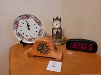 Photo showing four clock-related items on wooden surface against wall: vintage wooden mantle clock with floral motif, brass clock without glass dome and with ornamental top, floral patterned oval clock plate with Roman numerals, black digital clock with large red number display showing 12:03.