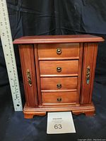 Front view of wooden jewelry box with four drawers and side doors closed. Ruler next to it shows approximate size: about 12 inches tall.