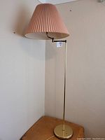 Full view of a tall brass floor lamp with a pleated rose-colored shade, shown against a wall and placed on a small wooden table.