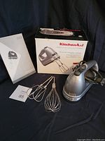 Photo of KitchenAid hand mixer in silver with two beaters, whisk attachments, original box and instruction manual on black background.