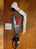 Photo showing two handheld electric massagers: one white with blue accents and one dark grey Conair model, both with cords coiled.