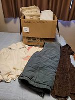 Box filled with various sweaters, vests, and sweatshirts, some displayed outside the box on a bed.