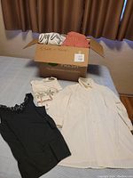 Photo of a box of assorted t-shirts and blouses laid on a table, showing four visible tops including a black lace sleeveless top, printed white t-shirt, white blouse, and orange ribbed top in the box.