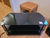 Front angled view showing the black smoked tempered glass shelves and metal cylindrical legs with a wooden stool in the background.