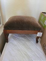 Full view of wooden foot stool with brown upholstered seat and carved legs.