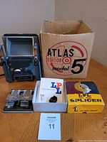 Photo showing Atlas Model 5 movie editor unit next to original box, LPL splicer box, and film splicing clamps with manual inside a white box.