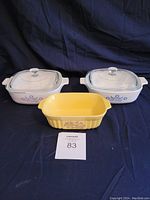 Photo showing two white square CorningWare casserole dishes with lids and one vintage yellow dish in front, all on dark fabric background.