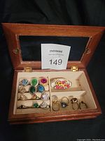 Open wooden jewelry case showing beige lining and 15 costume rings inside with varied designs including colored stones and floral enamel.