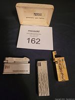 Front view of three metal lighters and Bentley Butane lighter case box showing the lid open with logo inside.