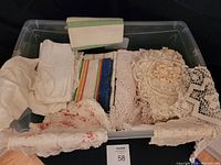 Clear plastic tote with assorted vintage linens including hand-sewn tablecloths, crocheted doilies, napkins, and kitchen towels in various colors and patterns.