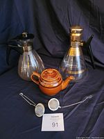 Two glass tea or coffee carafes with gold stripes and black handles, one Brown Betty brown ceramic teapot, and two metal mesh loose tea holders placed on a black cloth background.