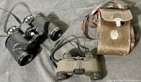 Two vintage binoculars placed side by side with brown carrying case; black textured binoculars and smaller greenish pair visible with straps.