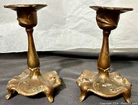 Pair of gilded metal candle holders in Art Nouveau style with ornate foot bases, photographed from front angle showing candle cups and bases.